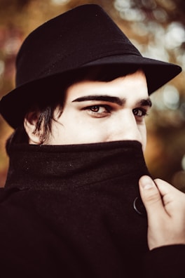 A person is wearing a black fedora and a coat with the collar pulled up, covering most of their lower face. The focus is on their eyes, which convey a mysterious or secretive expression. The background features a soft bokeh effect with warm tones of brown and green.
