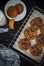 A set of freshly baked chocolate chip cookies are placed on a cooling rack lined with parchment paper. A plate with additional cookies and a bite taken out of one sits nearby, along with a glass of milk. A blue plaid cloth is positioned on the dark surface, with scattered crumbs and salt.
