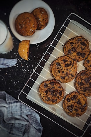 A set of freshly baked chocolate chip cookies are placed on a cooling rack lined with parchment paper. A plate with additional cookies and a bite taken out of one sits nearby, along with a glass of milk. A blue plaid cloth is positioned on the dark surface, with scattered crumbs and salt.