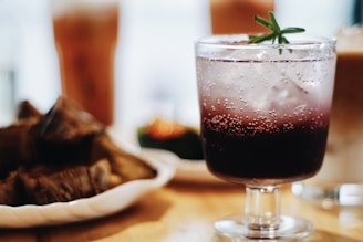 A close-up view of a fizzy, purple-hued beverage in a clear glass adorned with a sprig of rosemary. The background features blurred elements of food on plates, suggesting a dining setup in a warm atmosphere.