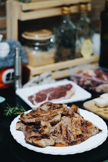 A plate of grilled meat is placed on a dark countertop, with other raw steaks and kitchen items in the background. Oil bottles and spices are arranged on a wooden rack, creating a cozy kitchen atmosphere.