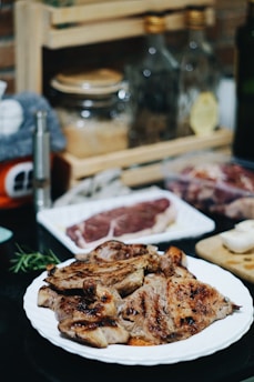 A plate of grilled meat is placed on a dark countertop, with other raw steaks and kitchen items in the background. Oil bottles and spices are arranged on a wooden rack, creating a cozy kitchen atmosphere.