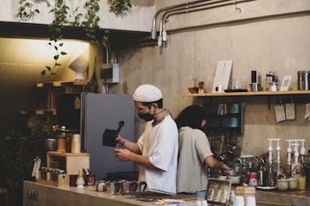 A cozy, rustic coffee shop with wooden shelves filled with jars and cups. Two people are making coffee, one wearing a white cap and mask, holding a coffee pot. Various plants hang from above, adding to the warm ambiance.