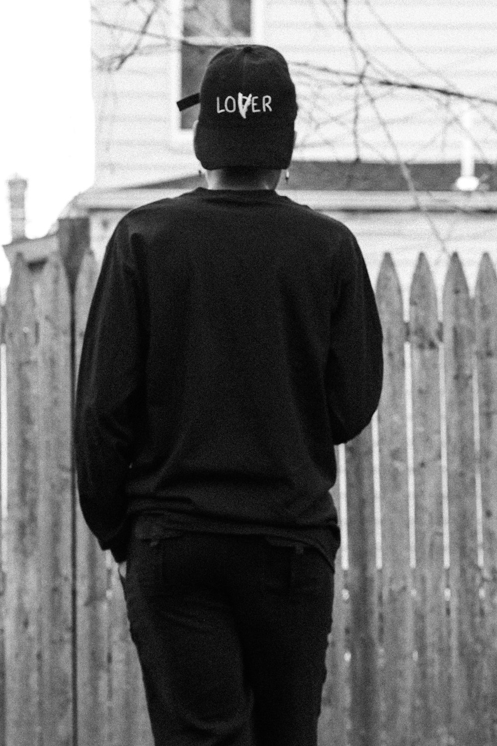 黒い長袖シャツと黒いズボンを着た男が白い木の柵の前に立つ