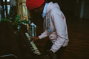 A person wearing a red beanie and a light-colored hoodie is playing an old, worn piano. The setting appears to be a dimly lit room with wooden floors and some decorative greenery and a white lantern nearby.