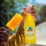 A hand holds a bottle of Jarritos pineapple soda alongside a popsicle coated with chili powder. The yellow-golden colors stand out against a blurred natural background with trees.