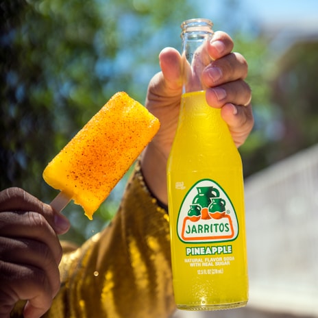 A hand holds a bottle of Jarritos pineapple soda alongside a popsicle coated with chili powder. The yellow-golden colors stand out against a blurred natural background with trees.