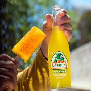 A hand holds a bottle of Jarritos pineapple soda alongside a popsicle coated with chili powder. The yellow-golden colors stand out against a blurred natural background with trees.