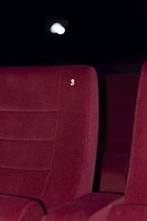 A red upholstered theater seat is prominently featured with the number three embossed on its backrest. The background is dimly lit, suggesting a cinema or theater environment.