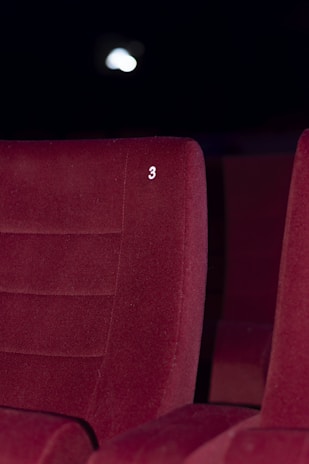 A red upholstered theater seat is prominently featured with the number three embossed on its backrest. The background is dimly lit, suggesting a cinema or theater environment.