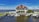 A large house with a red roof is situated beside a body of water. In front of the house, there are multiple docks with several boats moored. The sky is bright and partly cloudy, creating a peaceful and serene atmosphere.