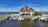 A large house with a red roof is situated beside a body of water. In front of the house, there are multiple docks with several boats moored. The sky is bright and partly cloudy, creating a peaceful and serene atmosphere.
