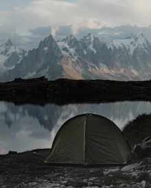 A green tent is pitched near a tranquil lake with a backdrop of towering, rugged mountains covered in patches of snow. The scene is peaceful, with the reflection of the mountains visible in the calm water. The sky is overcast with clouds, adding a sense of tranquility and solitude.
