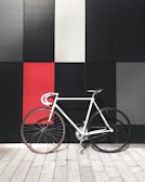 A white minimalist bicycle with a black wheel and a tan wheel is positioned against a geometric wall composed of large black, gray, and red panels. The floor consists of light gray paving stones.
