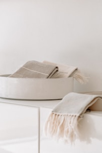 A white ceramic bowl containing folded beige and cream-colored towels. Another towel with fringed edges is draped over the edge of a minimalist white countertop.