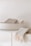 A white ceramic bowl containing folded beige and cream-colored towels. Another towel with fringed edges is draped over the edge of a minimalist white countertop.