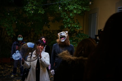 A group of people is gathered outdoors, surrounded by greenery. One person is wearing a realistic lion mask and suit, while another holds a transparent sphere. The background features dense foliage and outdoor lighting. The atmosphere is somewhat theatrical or unconventional.