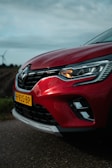 A close-up view of the front portion of a red car with a visible headlight and a license plate labeled H-935-BP. The car is parked on a road with a blurred background of fields and a wind turbine, under a cloudy sky.