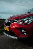 A close-up view of the front portion of a red car with a visible headlight and a license plate labeled H-935-BP. The car is parked on a road with a blurred background of fields and a wind turbine, under a cloudy sky.
