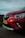 A close-up view of the front portion of a red car with a visible headlight and a license plate labeled H-935-BP. The car is parked on a road with a blurred background of fields and a wind turbine, under a cloudy sky.