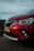A close-up view of the front portion of a red car with a visible headlight and a license plate labeled H-935-BP. The car is parked on a road with a blurred background of fields and a wind turbine, under a cloudy sky.