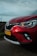 A close-up view of the front portion of a red car with a visible headlight and a license plate labeled H-935-BP. The car is parked on a road with a blurred background of fields and a wind turbine, under a cloudy sky.