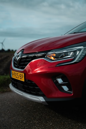 A close-up view of the front portion of a red car with a visible headlight and a license plate labeled H-935-BP. The car is parked on a road with a blurred background of fields and a wind turbine, under a cloudy sky.
