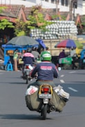 A motorcyclist wearing a Go-Jek helmet and a jacket rides along a busy street with bags strapped to the sides of the motorcycle. In the background, there's a cart with an umbrella, a small stall, and a truck carrying many stacked items. Several people are visible in the scene, engaged in various activities.