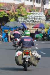 A motorcyclist wearing a Go-Jek helmet and a jacket rides along a busy street with bags strapped to the sides of the motorcycle. In the background, there's a cart with an umbrella, a small stall, and a truck carrying many stacked items. Several people are visible in the scene, engaged in various activities.