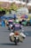 A motorcyclist wearing a Go-Jek helmet and a jacket rides along a busy street with bags strapped to the sides of the motorcycle. In the background, there's a cart with an umbrella, a small stall, and a truck carrying many stacked items. Several people are visible in the scene, engaged in various activities.