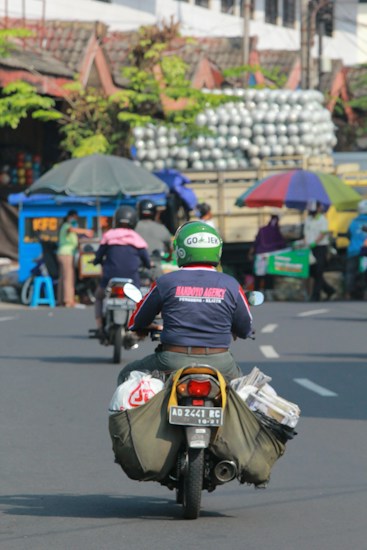 A motorcyclist wearing a Go-Jek helmet and a jacket rides along a busy street with bags strapped to the sides of the motorcycle. In the background, there's a cart with an umbrella, a small stall, and a truck carrying many stacked items. Several people are visible in the scene, engaged in various activities.