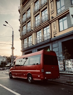 A red van is parked on a city street near a tall building with multiple windows and decorative balconies. The sky is overcast with hints of sunlight peeking through, adding a subtle glow. An urban setting with streetlights, power lines, and road signs gives a vibe of early morning or late afternoon.