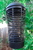 A large black electronic insect zapper is suspended among lush green leaves. The cylindrical device has a grid-like structure designed to attract and eliminate insects, with remnants visible within its confines. Sunlight filters through the leaves, creating a dappled effect around the device.