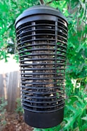 A large black electronic insect zapper is suspended among lush green leaves. The cylindrical device has a grid-like structure designed to attract and eliminate insects, with remnants visible within its confines. Sunlight filters through the leaves, creating a dappled effect around the device.