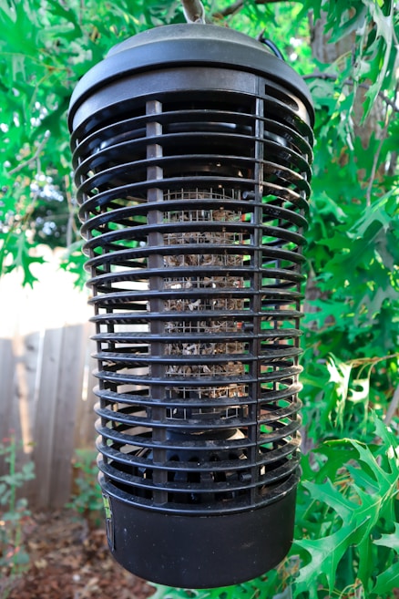 A large black electronic insect zapper is suspended among lush green leaves. The cylindrical device has a grid-like structure designed to attract and eliminate insects, with remnants visible within its confines. Sunlight filters through the leaves, creating a dappled effect around the device.
