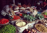 A vibrant market scene with a variety of fresh produce displayed in woven baskets. The produce includes red chili peppers, pineapples, ginger, onions, garlic, and various other fruits and vegetables. A woman is handling the peppers, while a few other people are seen in the background.