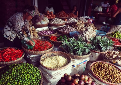 A vibrant market scene with a variety of fresh produce displayed in woven baskets. The produce includes red chili peppers, pineapples, ginger, onions, garlic, and various other fruits and vegetables. A woman is handling the peppers, while a few other people are seen in the background.