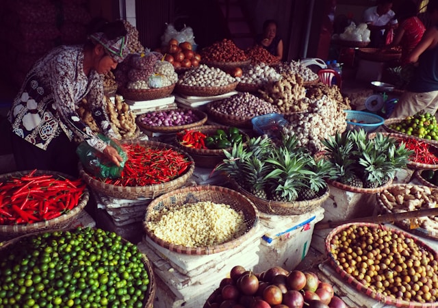 A vibrant market scene with a variety of fresh produce displayed in woven baskets. The produce includes red chili peppers, pineapples, ginger, onions, garlic, and various other fruits and vegetables. A woman is handling the peppers, while a few other people are seen in the background.