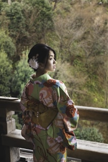 A person wearing a traditional Japanese kimono stands on a wooden balcony, looking into the distance. The kimono features a colorful floral pattern with shades of green, pink, and orange. A large bow, or obi, is tied at the back. The hairstyle is adorned with white flowers and hairpins. Lush greenery and trees provide a natural backdrop.