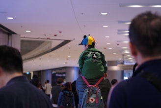 A child wearing a colorful cap and a green jacket with a bear design is sitting on an adult's shoulders in a busy indoor setting, likely a shopping mall or an airport. The scene includes several other people walking in the background, and the lighting gives a warm ambiance.