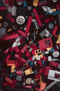 A chaotic pile of colorful LEGO pieces in various shapes and sizes, including red, yellow, blue, and black blocks as well as wheels and windows.
