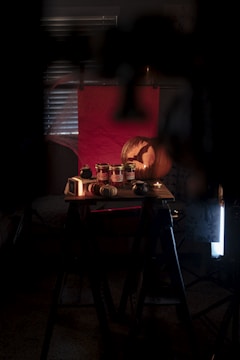 A dimly lit room with jars of preserved food on a wooden table, accompanied by small decorative pumpkins and gourds. A large carved pumpkin with a silhouette design is illuminated in the background. There is a red backdrop and a window with closed blinds partially visible.