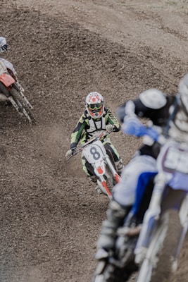 A group of motocross riders navigating a dirt track. The rider in focus is wearing a green and black outfit and a helmet with goggles, riding a bike with the number 8. The track is rough with visible gravel and dirt, adding to the intensity of the scene.
