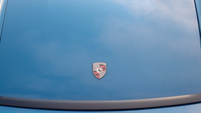 A car hood is painted in a blue color with a Porsche logo prominently displayed in the center.
