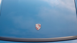 A car hood is painted in a blue color with a Porsche logo prominently displayed in the center.