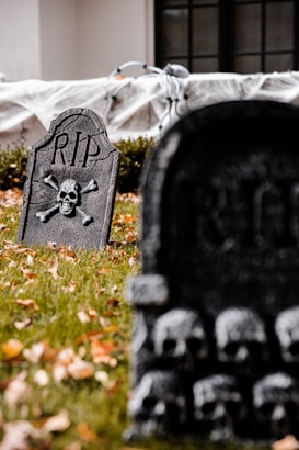 Tombstones with skull and crossbones designs are placed on a grassy lawn, surrounded by fallen autumn leaves. A white sheet or decoration is visible in the background near the house.