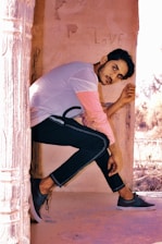 A person sits pensively against a light-colored wall with the word 'love' faintly visible behind them. They are wearing a casual outfit with a light-colored shirt and dark pants, accompanied by sneakers. The scene has a warm, rustic aesthetic with stone pillars and outdoor elements visible.