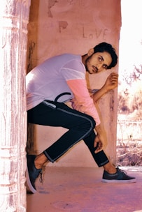 A person sits pensively against a light-colored wall with the word 'love' faintly visible behind them. They are wearing a casual outfit with a light-colored shirt and dark pants, accompanied by sneakers. The scene has a warm, rustic aesthetic with stone pillars and outdoor elements visible.