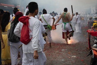 A group of people dressed in traditional white clothing participate in a vibrant street procession. One participant is energetically jumping over a small explosive device that emits smoke and sparks. The atmosphere is festive, with tables decorated with yellow flowers and other offerings.