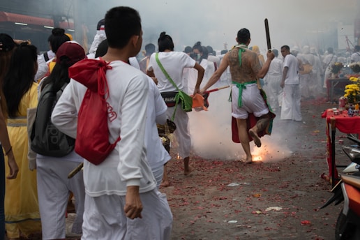 A group of people dressed in traditional white clothing participate in a vibrant street procession. One participant is energetically jumping over a small explosive device that emits smoke and sparks. The atmosphere is festive, with tables decorated with yellow flowers and other offerings.